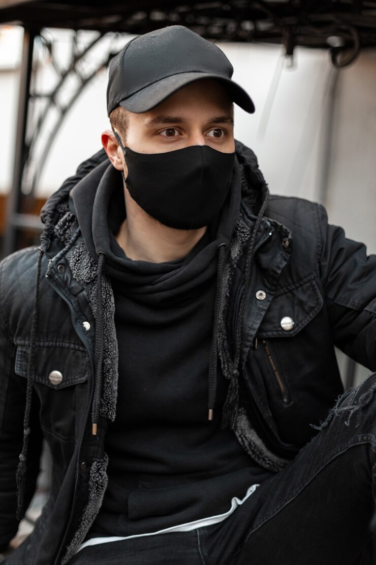 Foto elegante jovem bonito com uma máscara médica e um boné preto em uma jaqueta preta da moda com um capuz sentado na rua. estilo masculino urbano legal e conceito covid-19