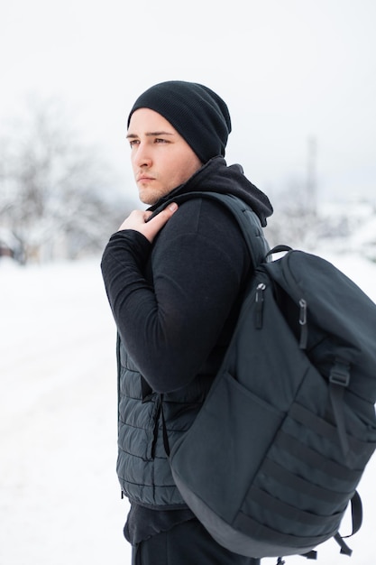 Elegante jovem bonito em agasalhos de inverno preto de moda com uma mochila viaja em um dia de inverno. Cara modelo em roupas pretas viaja em tempo de neve