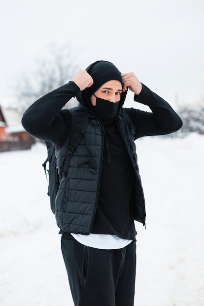 Elegante jovem bonito em roupas de inverno preto da moda e uma máscara protetora com uma mochila coloca um capuz ao ar livre com neve.