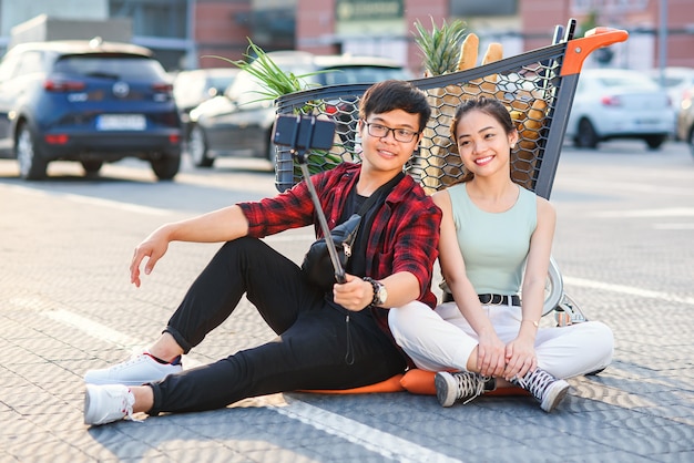 Elegante jovem casal vietnamita sentado no chão perto de carrinho de compras e fazendo selfie foto juntos.