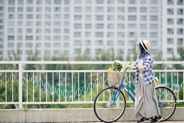 Elegante jovem chinesa com camisa xadrez e calças largas em pé na ponte ao lado de sua bicicleta e apreciando a vista