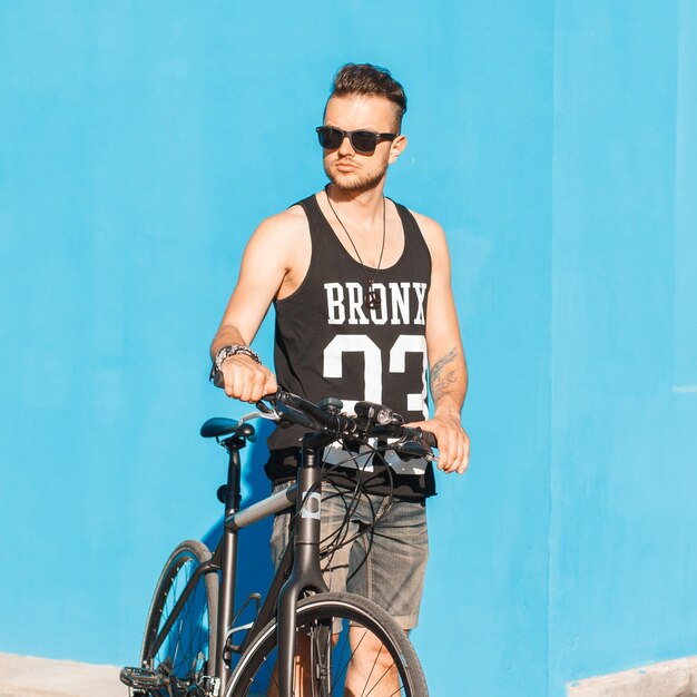 Elegante jovem ciclista hipster com óculos de sol de moda em roupas de verão com uma bicicleta em um fundo azul na rua