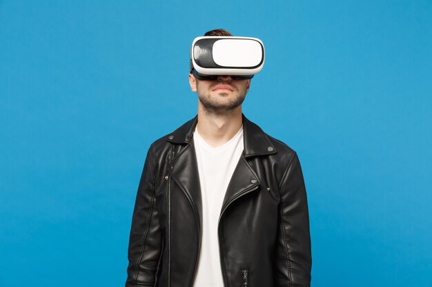 Elegante jovem com barba por fazer em t-shirt branca de jaqueta preta olhando no fone de ouvido, realidade virtual vr isolada no retrato de estúdio de fundo de parede azul. Conceito de estilo de vida de emoções de pessoas. Simule o espaço da cópia.