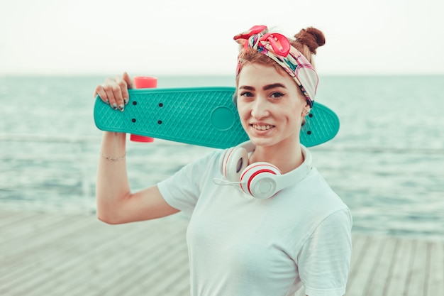 Foto elegante jovem com uma bandana na cabeça, fones de ouvido com skate nas mãos, sorrindo na praia.