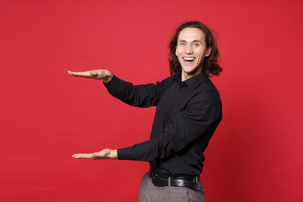 Elegante jovem de cabelos compridos encaracolado na camisa preta posando isolado no retrato de estúdio de fundo de parede vermelha. Conceito de estilo de vida de emoções sinceras de pessoas. Mock up espaço de cópia. Gesticulando demonstrando tamanho.