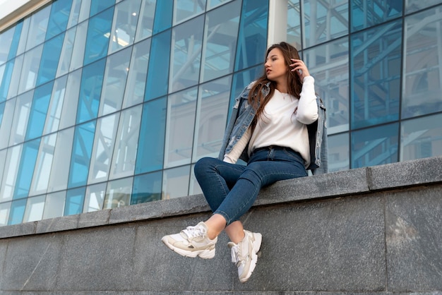 Elegante jovem descansando na rua da cidade de verão Linda estudante em jeans sentada perto de um prédio moderno