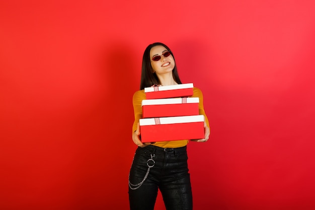Elegante jovem feliz e sorridente em óculos de sol vermelhos segura três caixa de presente vermelho-branca isolada na superfície vermelha do estúdio.