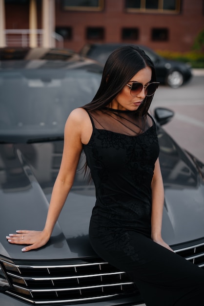 Elegante jovem fica perto do carro em um vestido preto. moda e estilo de negócios