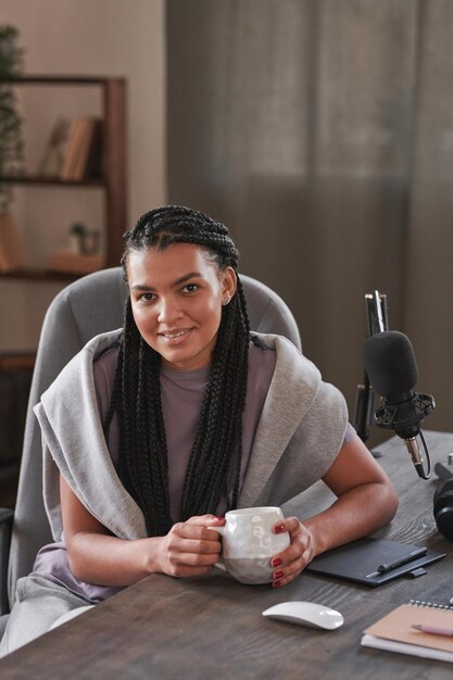 Elegante jovem influenciadora sentada à mesa na sala de loft tendo pausa bebendo chá sorrindo para ca