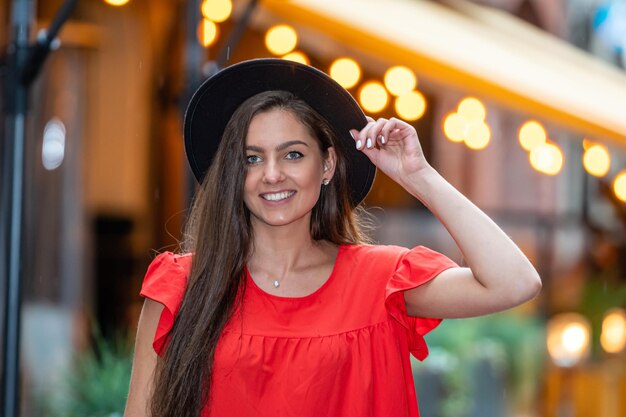 Foto elegante jovem sensual mulher em um vestido vermelho e um chapéu preto em um fundo de luzes de café de rua