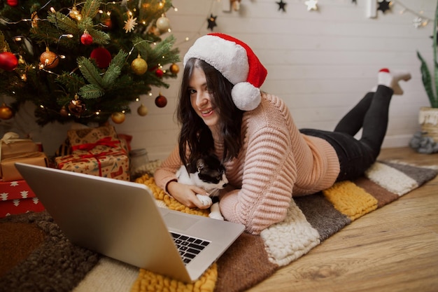 Elegante jovem sentada com gato bonito no laptop no tapete debaixo da árvore de natal com presentes Garota feliz com chapéu de Papai Noel trabalhando ou fazendo compras online com gatinho no quarto moderno