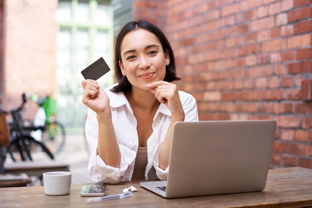 Elegante joven asiática haciendo compras en línea sentada con tarjeta de crédito y computadora portátil comprando en Internet y pagando sin contacto con las facturas