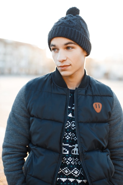 Elegante joven atractivo con un sombrero de moda en un suéter de punto azul vintage en una chaqueta de invierno cálida de moda está