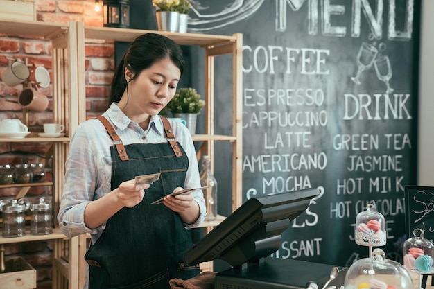 elegante joven dueña de un café usa delantal de pie en el mostrador contando el dinero que ganan los clientes hoy. dama china asiática barista contabilidad finanzas en cafetería. concepto exitoso de negocio de inicio