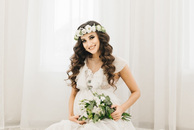 Elegante joven embarazada en ropa blanca con rizos y flores. Fotos esperando bebé