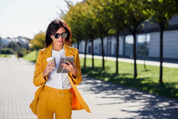 Elegante joven empresaria utiliza tablet PC mientras camina con una taza de café