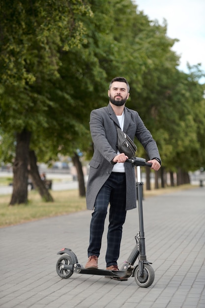 Elegante joven empresario contemporáneo de pie en scooter eléctrico en el entorno urbano contra la hilera de árboles verdes que crecen a lo largo de la carretera