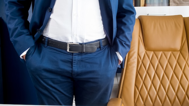 Foto elegante joven empresario en traje azul de pie en la oficina moderna con sillón de cuero marrón