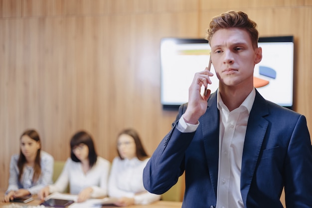 Foto elegante joven empresario vistiendo una chaqueta y una camisa en el fondo de una oficina de trabajo con personas hablando por un teléfono móvil