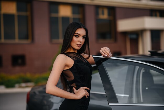 Elegante joven se encuentra cerca del coche en un vestido negro. Moda y estilo empresarial