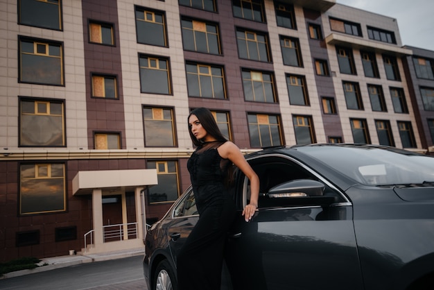 Foto elegante joven se encuentra cerca del coche con un vestido negro. moda y estilo empresarial