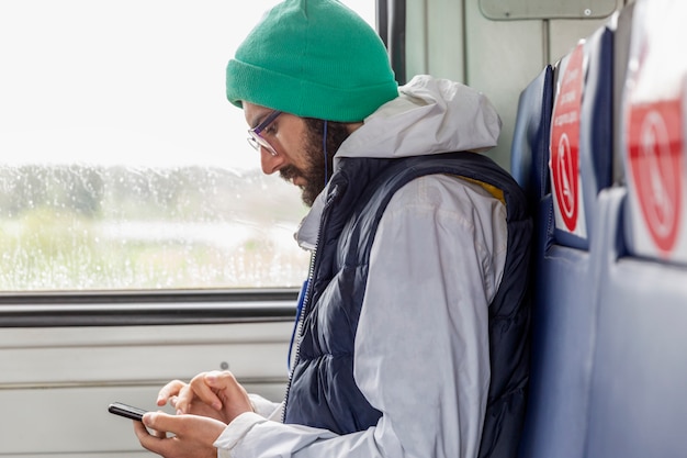 Elegante joven con gafas se sienta en un vagón de tren con marcas para asientos de pasajeros y mira en un teléfono inteligente.