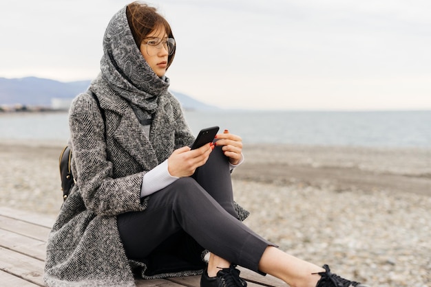 Elegante joven morena con gafas y un abrigo gris sentado en la playa junto al mar en tiempo nublado