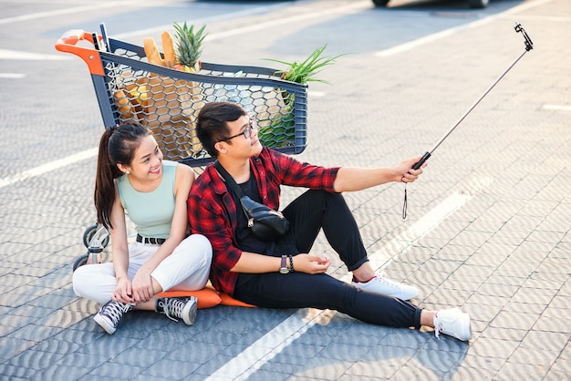 Elegante joven pareja vietnamita sentado en el suelo cerca del carrito de la compra y haciendo selfie foto juntos.