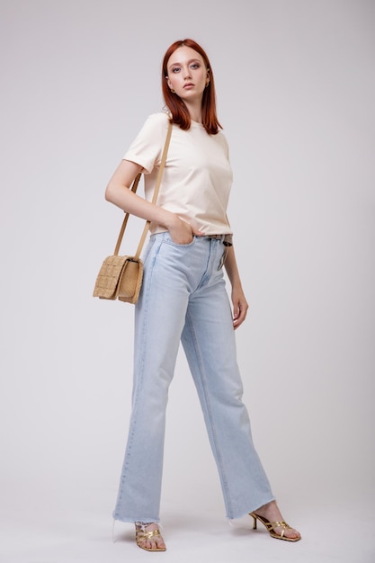 Elegante joven pelirroja con bonitos jeans azules, camiseta, bolso posando sobre fondo blanco