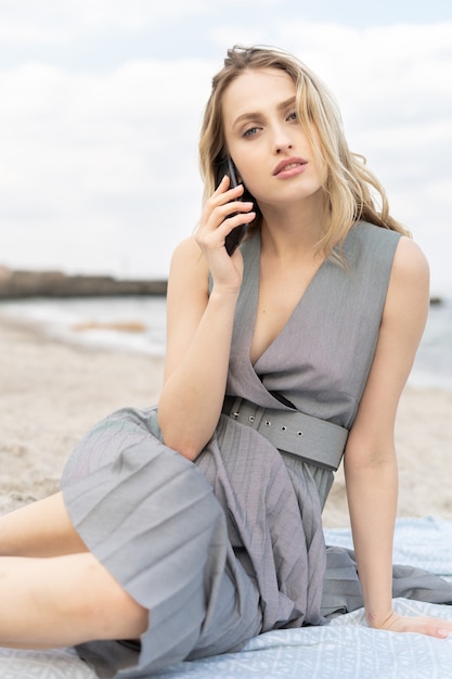 Elegante joven rubia está hablando por teléfono móvil en una playa disfrutando del clima