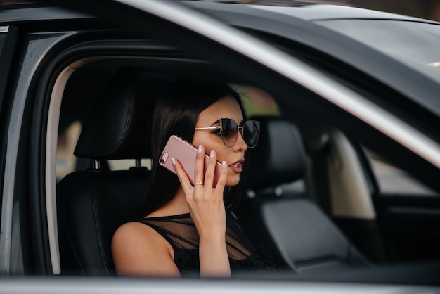 Elegante joven sentada en un coche de clase ejecutiva en un vestido negro y hablando por teléfono. moda y estilo empresarial.