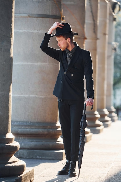 Elegante joven con sombrero negro y ropa con paraguas en las manos al aire libre en la ciudad