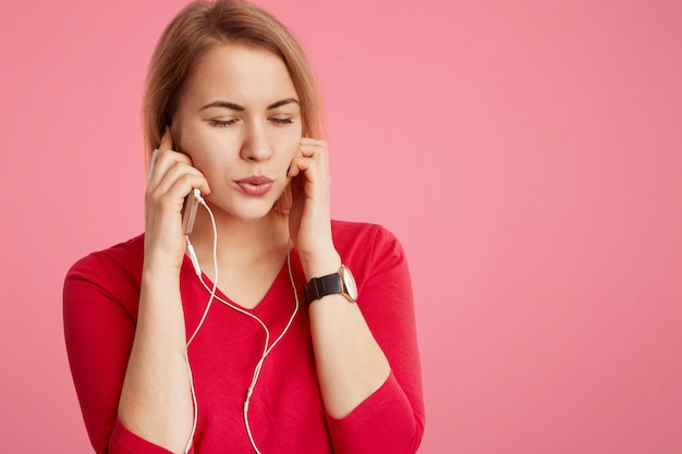 Elegante joven usa un teléfono celular moderno y auriculares para escuchar su música favorita o pista de la lista de reproducción, cierra los ojos mientras siente relajación y gran placer, aislado en rosa