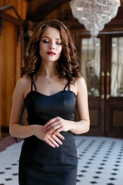Elegante junge Frau im schwarzen Abendkleid, das im Weinleseinnenraum aufwirft. Modeschuss.