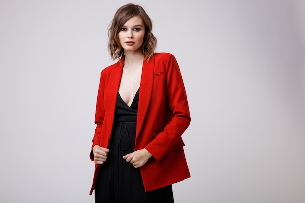Elegante junge Frau im schwarzen Abendkleid mit tiefem Ausschnitt, roter Jacke auf weißem Hintergrund