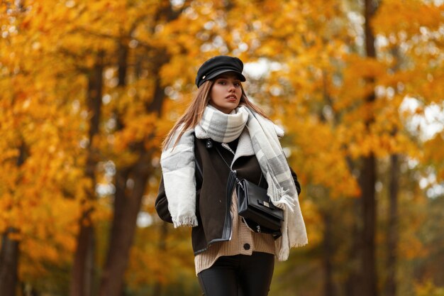 Elegante junge Frau in einem schicken Hut in einer stilvollen braunen Jacke mit einer Ledertasche mit einem Schal, der auf dem Hintergrund von Bäumen mit goldenen Blättern im Park an einem Herbsttag aufwirft. Schönes Mädchen im Freien.