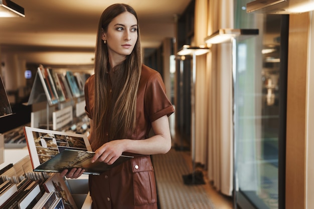 Elegante junge verführerische Frau, die im Vintage-Buchladen einkauft, Magazin hält, sich umdrehen Blick außerhalb Fenster.