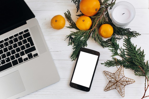 Elegante laptop de natal plano e telefone com tela vazia em fundo branco de madeira com ornamentos sazonais de laranjas e conceito de publicidade de estrela dourada