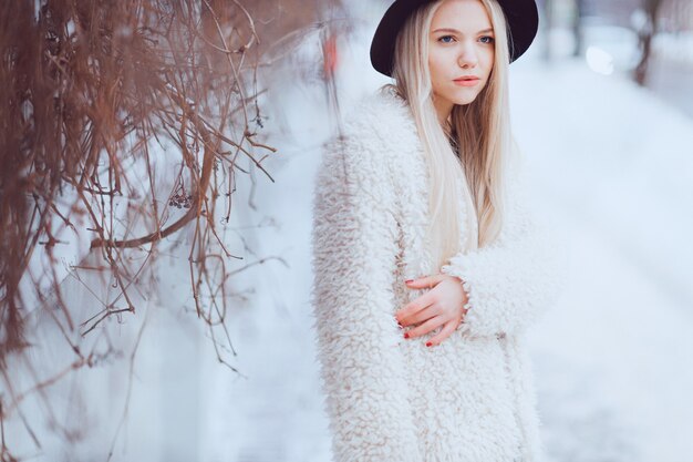 Elegante linda garota loira de casaco e chapéu posando perto da cerca na rua