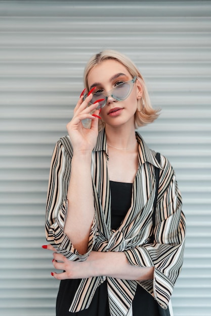 Elegante linda jovem com um corte de cabelo curto e grandes manicures vermelhas em roupas da moda com uma bolsa usando óculos de sol de verão legal