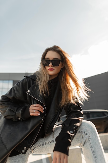 Elegante linda jovem em roupas da moda com óculos de jaqueta de couro com capuz jeans e poses de bolsa na cidade em dia ensolarado