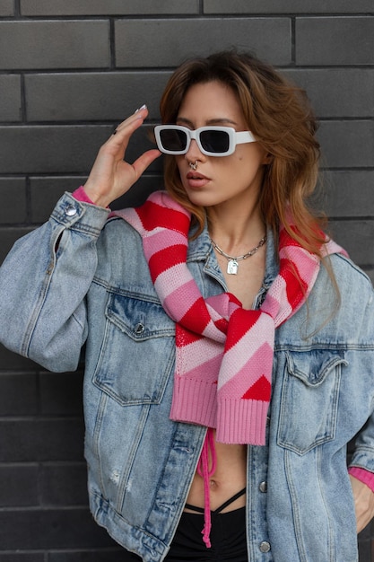 Elegante linda jovem ruiva hipster modelo em roupas urbanas da moda com jeans um moletom rosa e um top está usando óculos escuros modernos e de pé perto de uma parede de tijolos pretos na cidade