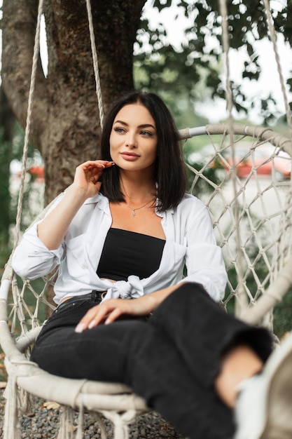 Elegante linda mulher de negócios bem-sucedida em uma elegante camisa branca e jeans preto senta e descansa em um balanço de malha no parque