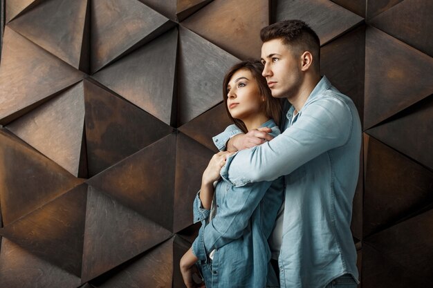 Elegante lindo casal jovem com roupas jeans azul perto de uma parede de madeira estampada