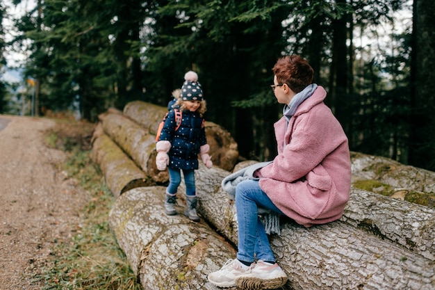 Elegante madre con corte de pelo corto se sienta en un tronco con su linda hija en un bosque