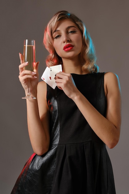 Elegante maquiagem brilhante feminina loira encaracolada em vestido preto elegante segurando duas cartas de baralho e taça de champanhe posando contra o fundo cinza do estúdio Jogo de poker closeup