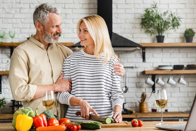 Elegante marido maduro abraçando a esposa enquanto ela cozinha salada na cozinha