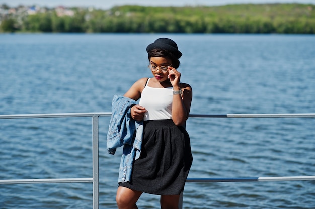 Elegante modelo afro-americano no chapéu de óculos, jaqueta jeans e saia preta posou ao ar livre no cais contra o lago.