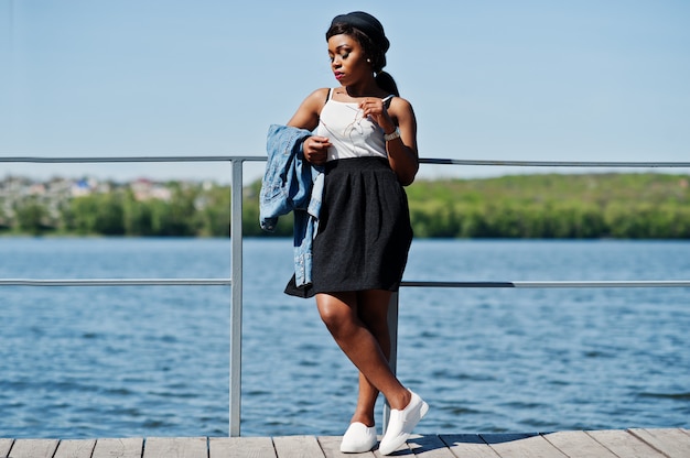 Elegante modelo afro-americano no chapéu de óculos, jaqueta jeans e saia preta posou ao ar livre no cais contra o lago.