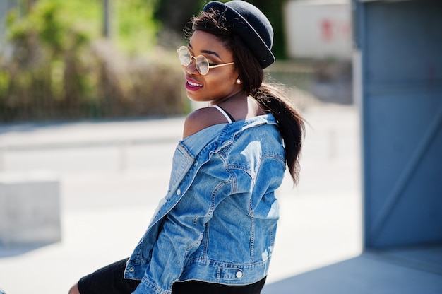 Elegante modelo afroamericano en gafas de sombrero, chaqueta de jeans y falda negra planteada al aire libre.